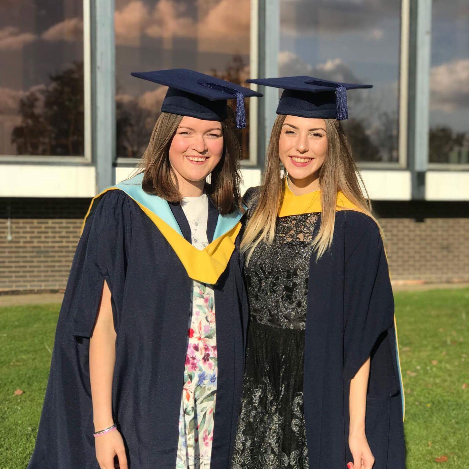 The Coffee & Jam Studios duo in graduation gowns and caps standing outdoors.